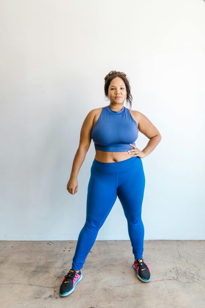A confident woman poses in blue activewear indoors, promoting body positivity and self-love.
